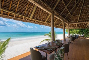 Un área de comedor frente a la playa, con una terraza de madera, mesas dispuestas para comer y vista al océano.