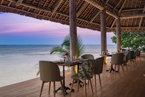 Un área de comedor junto al mar con mesas y sillas de madera, techo de paja y vistas al océano.