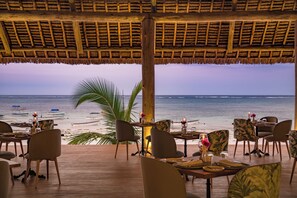 Un espace extérieur aménagé pour les repas, avec des tables et des chaises en bois, offrant une vue sur l’océan et les bateaux qui naviguent au loin.