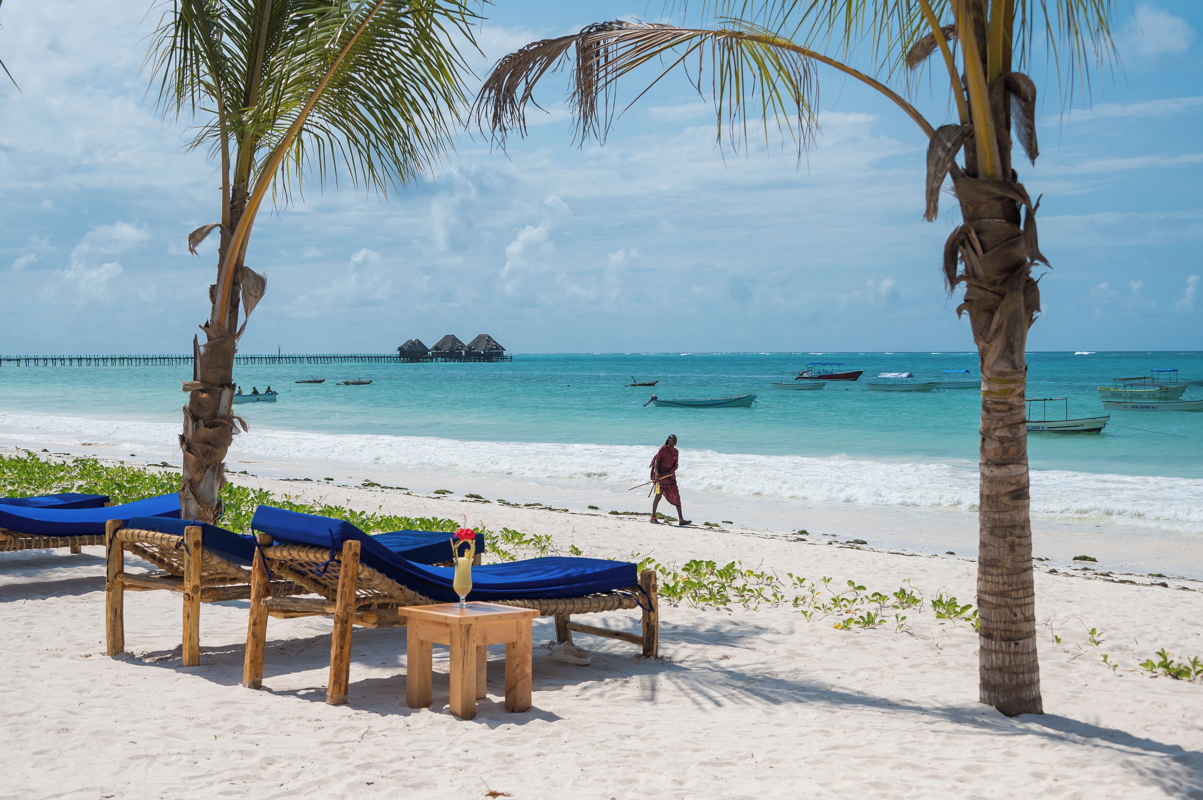 A beach with wooden lounge chairs and a small table with a drink, palm trees, and a person walking.