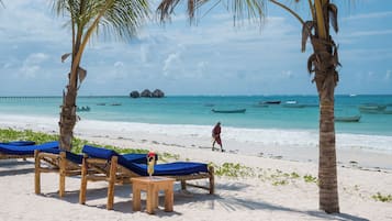 A beach with wooden lounge chairs and a small table with a drink, palm trees, and a person walking.