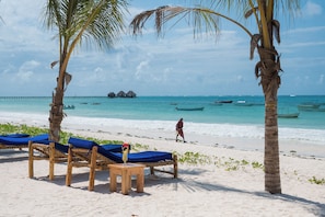 Ein Strand mit Holzliegen, einem kleinen Tisch mit einem Getränk, Palmen und einer Person, die spazieren geht.