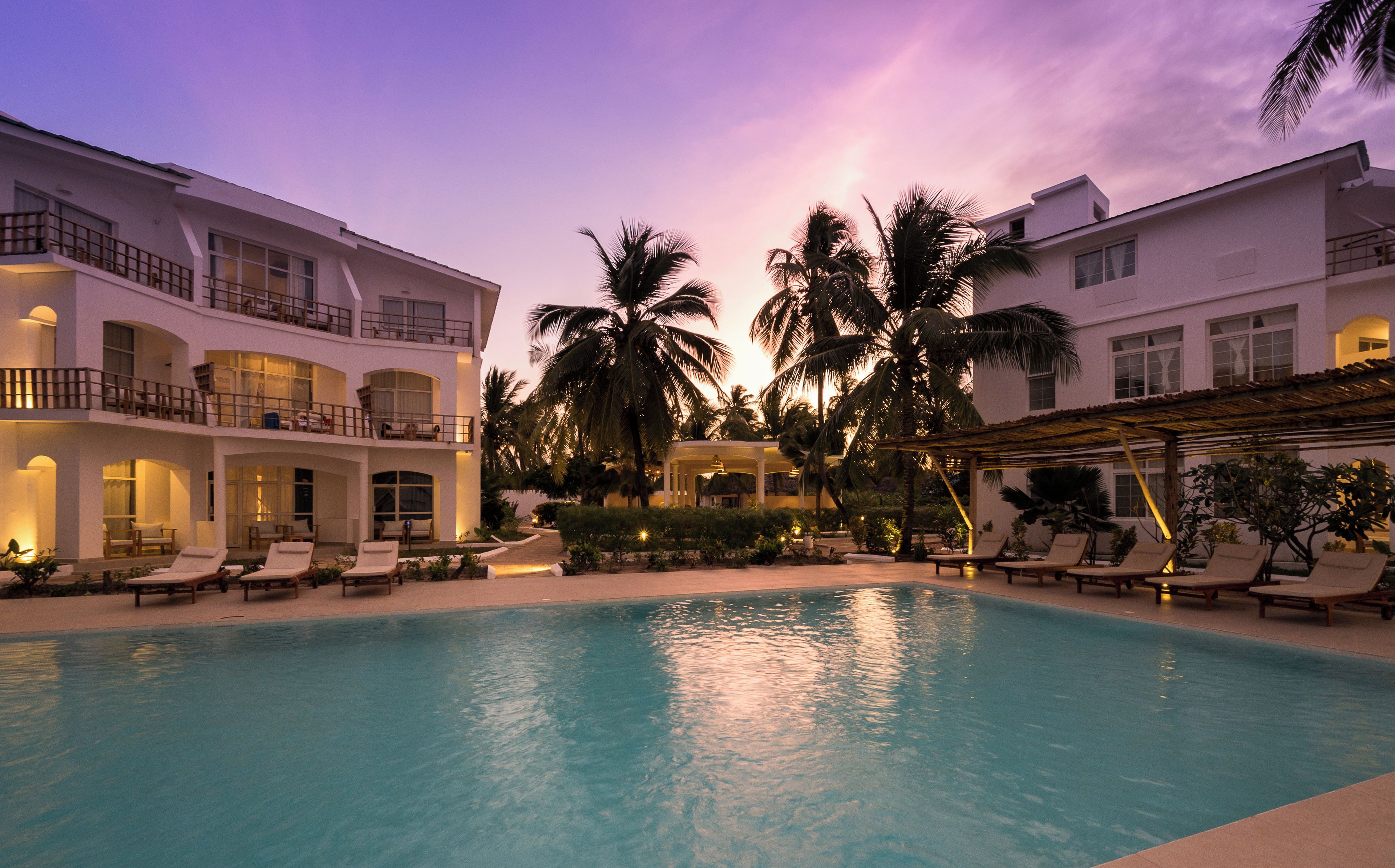 Zona de piscina en un resort con sillones de descanso y un espacio de estar cubierto, rodeada de palmeras y un edificio con varios balcones.