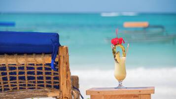 A wooden chair with a blue cushion and a glass of a tropical drink with a straw and a red garnish on a wooden table by the beach.