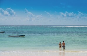 Una playa con agua turquesa cristalina, un bote pequeño y dos personas caminando por la orilla.