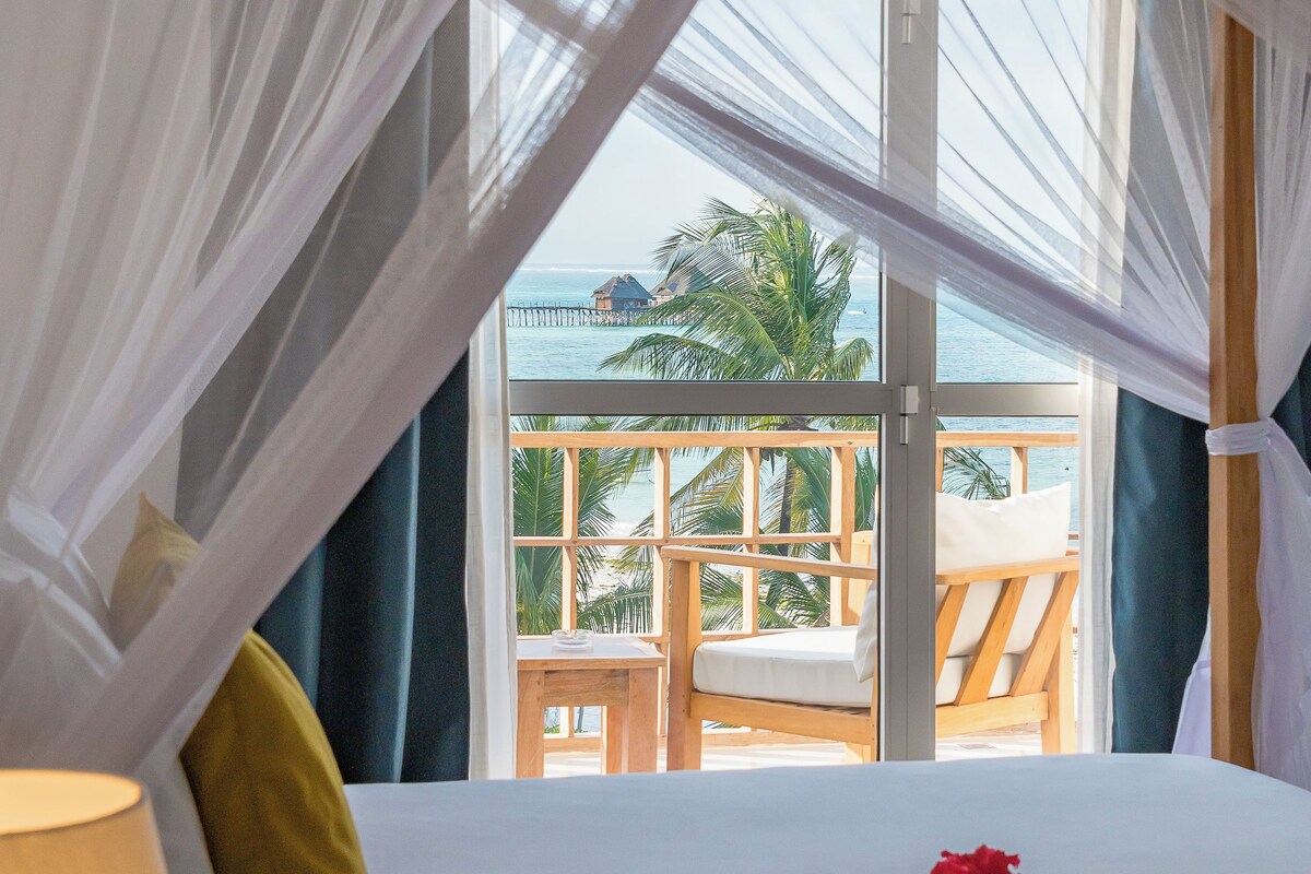 Ein Zimmer mit einem Bett, einem Fenster mit Blick auf einen Balkon und Palmen sowie einem Baldachin über dem Bett.