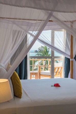Ein Zimmer mit einem Bett, einem Fenster mit Blick auf einen Balkon und Palmen sowie einem Baldachin über dem Bett.