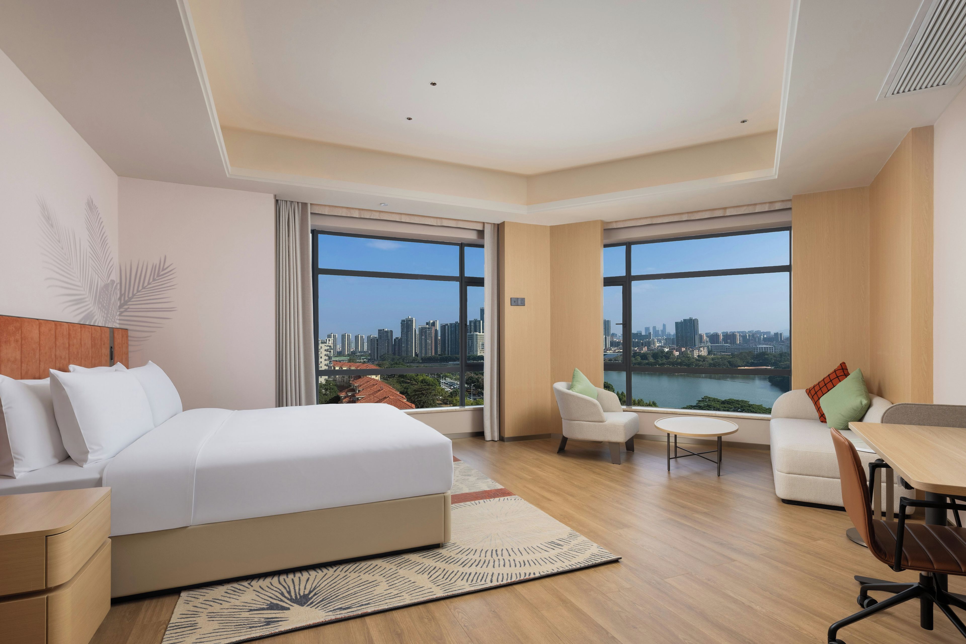 Ein modernes Hotelzimmer mit einem großen Bett, einem Schreibtisch, einem Sessel und zwei Fenstern mit Blick auf die Stadt.