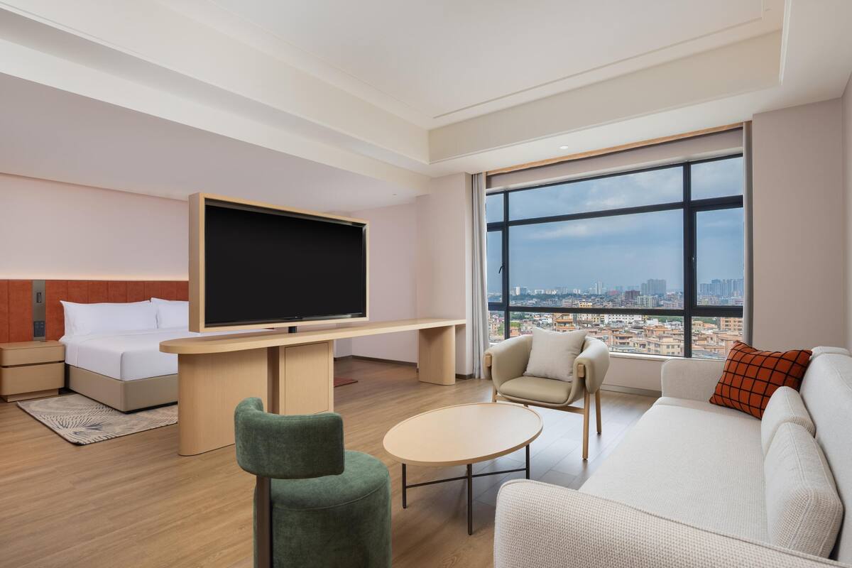 Une chambre d’hôtel moderne équipée d’un lit, d’une télévision, d’un bureau, d’un canapé et d’une grande fenêtre donnant sur la ville.