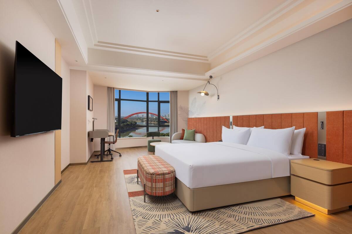 Une chambre d’hôtel dotée d’un grand lit, de tables de chevet, d’un téléviseur à écran plat, d’un bureau et d’une fenêtre donnant sur le paysage urbain.