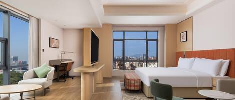 A modern hotel room with a large bed, a desk, a chair, a small table, and a view of the city through large windows.