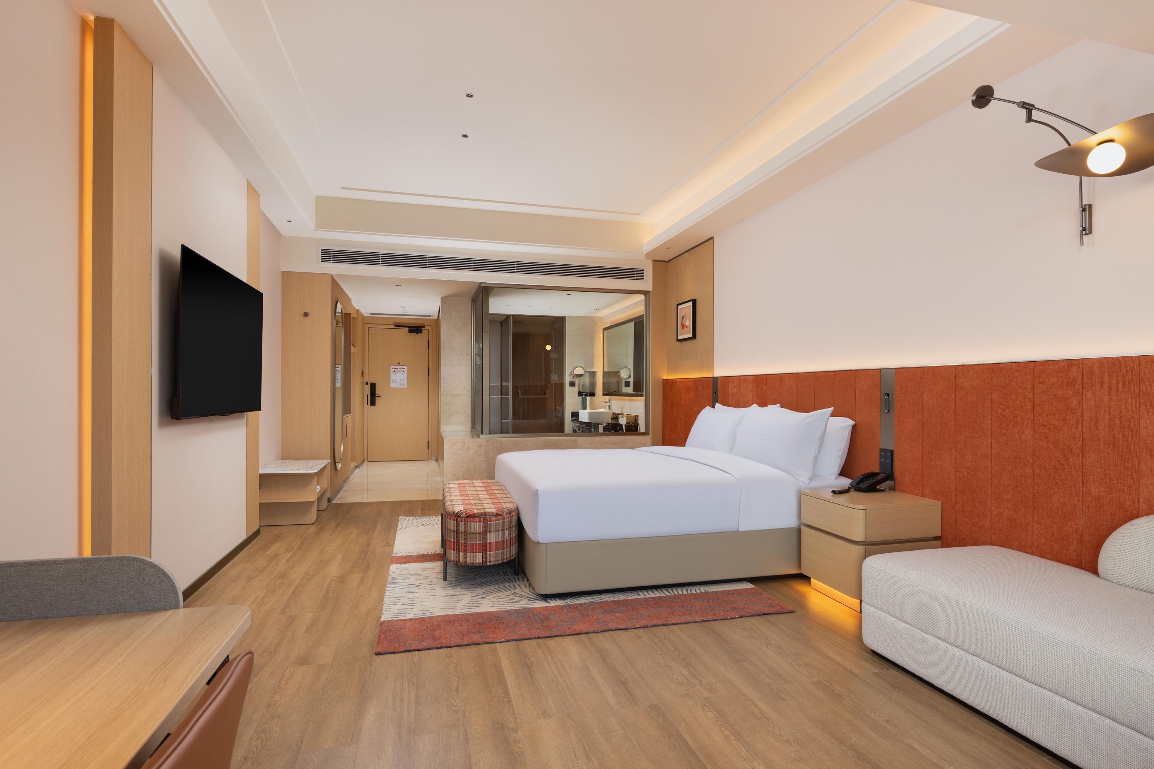 Ein modernes Hotelzimmer mit einem großen Bett, einem Nachttisch, einem kleinen Sitzbereich mit Sofa und Hocker sowie einem wandmontierten Flachbildfernseher.