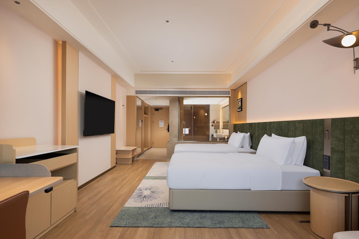 A hotel room with a bed, a desk, a TV, and a bathroom visible through a glass door.
