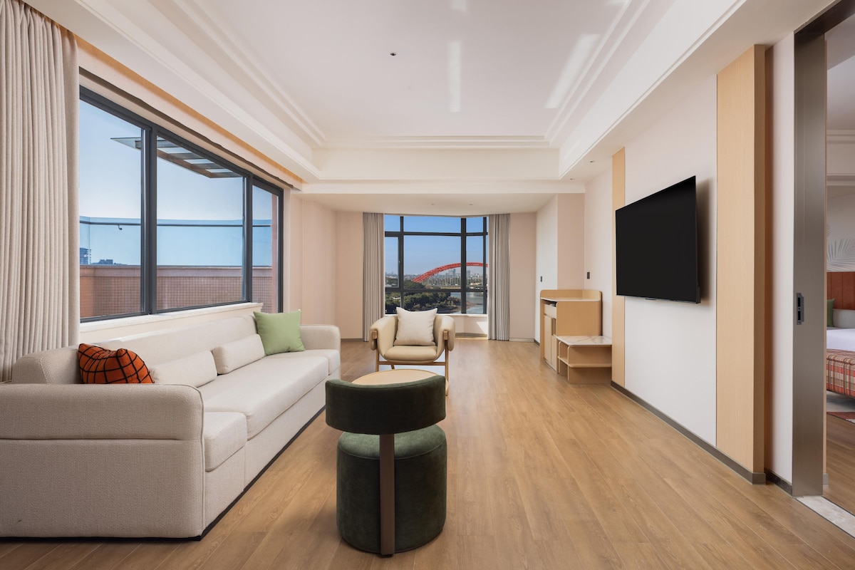 Une chambre d’hôtel moderne dotée d’une grande fenêtre, d’un canapé, d’un pouf rond, d’une télévision et d’un balcon avec une structure rouge en arrière-plan.