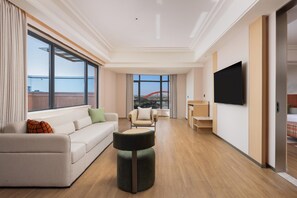 Une chambre d’hôtel moderne dotée d’une grande fenêtre, d’un canapé, d’un pouf rond, d’une télévision et d’un balcon avec une structure rouge en arrière-plan.