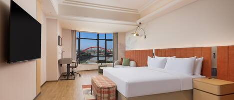 A hotel room with a large bed, bedside tables, a flat-screen TV, a desk, and a window with a cityscape view.