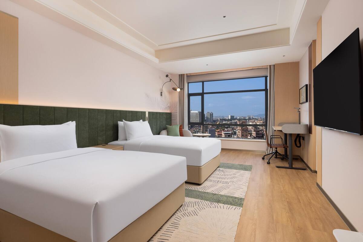 Une chambre d’hôtel avec deux lits, un bureau, une chaise, une télévision et une grande fenêtre donnant sur la ville.