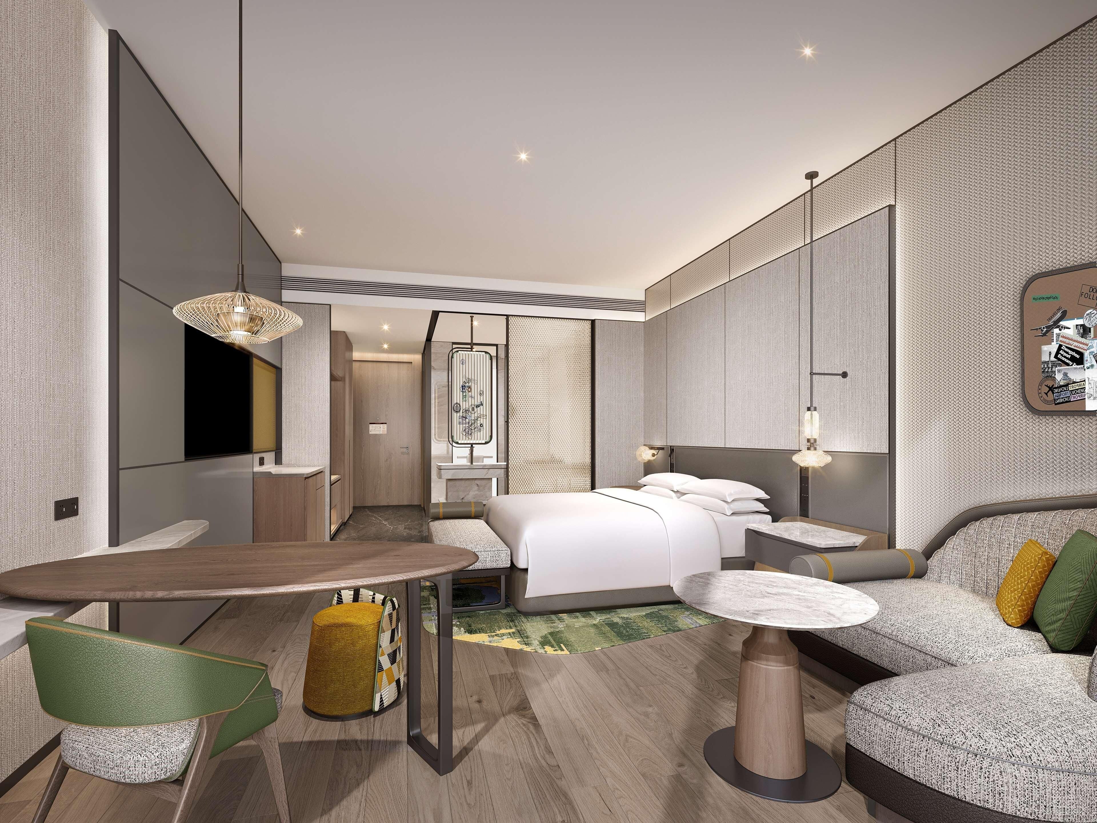A modern hotel room with a bed, a sofa, a dining table, and a small kitchen area.