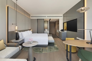 Quarto de hotel moderno com uma cama grande, mesas de cabeceira, uma mesa redonda, um sofá, uma TV e uma cozinha compacta.