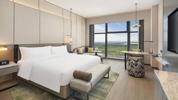 A modern hotel room with a large bed, bedside tables, a desk, a chair, a TV, and a view of the city.