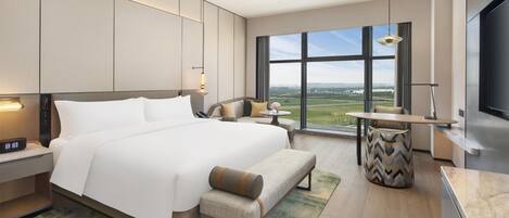 A modern hotel room with a large bed, bedside tables, a desk, a chair, a TV, and a view of the city.