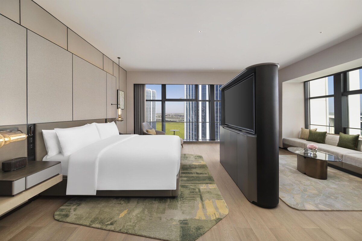 Une chambre d’hôtel moderne dotée d’un grand lit, d’un téléviseur à écran plat, d’un canapé et offrant une vue sur le paysage urbain grâce à de grandes fenêtres.