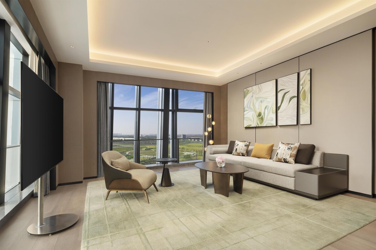Ein modernes Wohnzimmer mit einem großen Flachbildfernseher, einer Couch, einem Couchtisch und Blick auf eine Stadtlandschaft durch große Fenster.