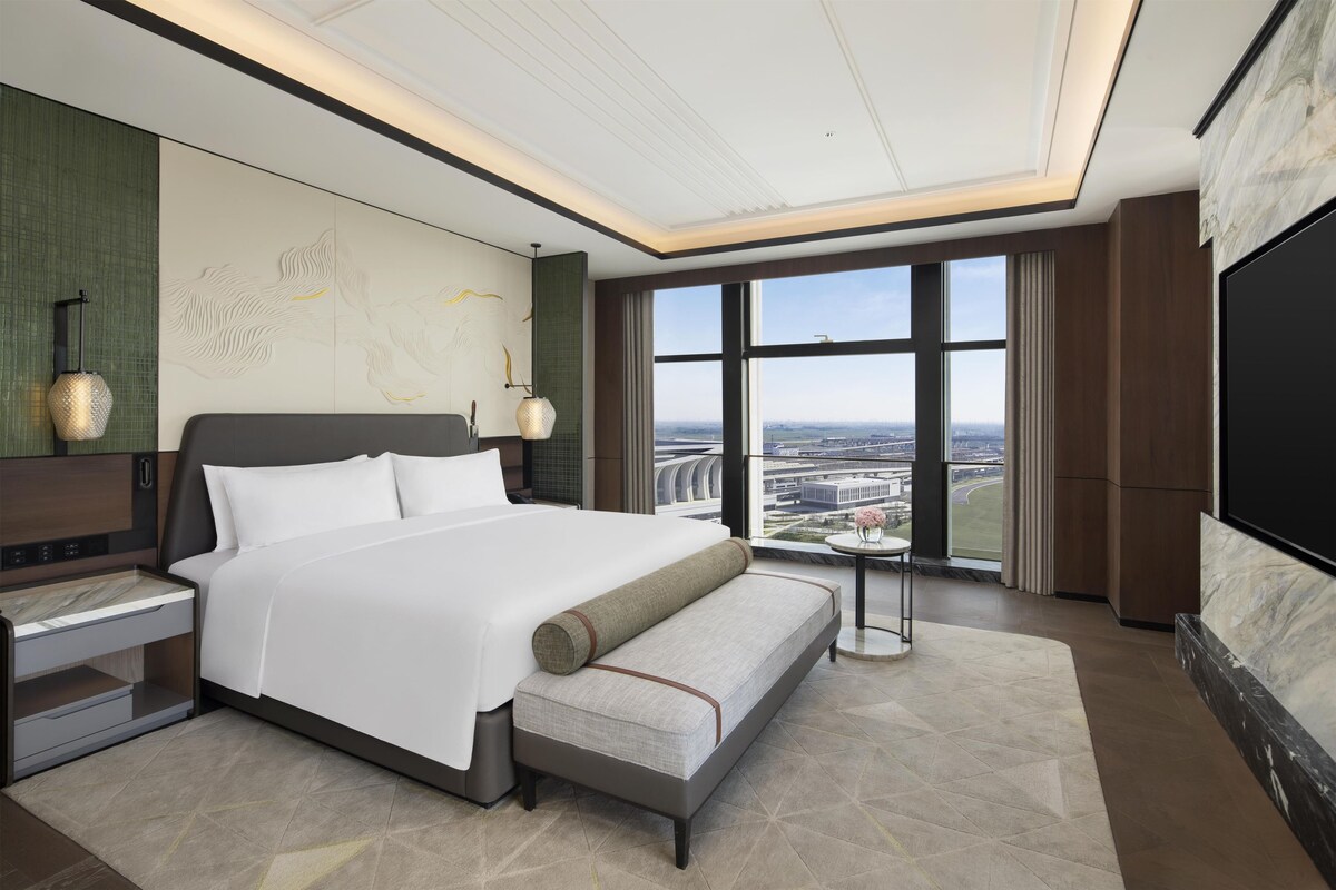 Ein modernes Hotelzimmer mit einem großen Bett, Nachttischen, einem Flachbildfernseher und Blick auf die Stadtlandschaft.