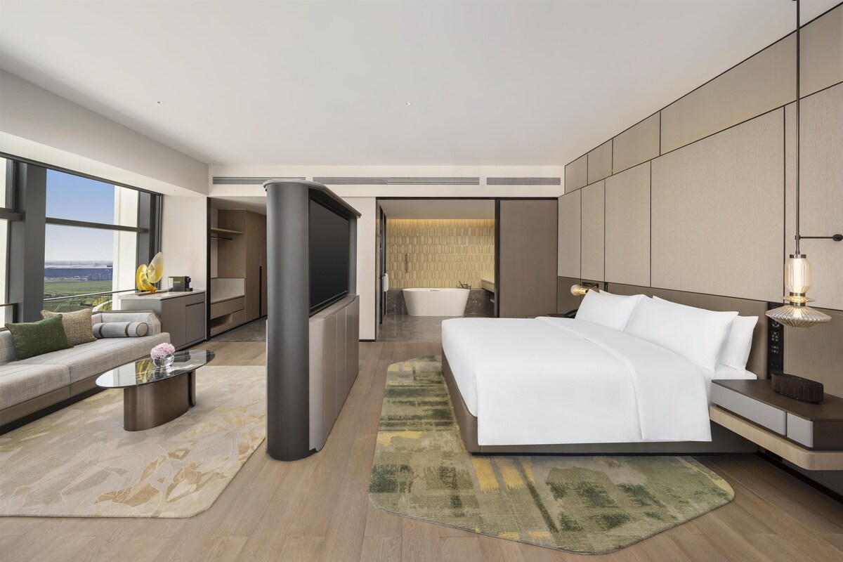 Ein modernes Hotelzimmer mit einem großen Bett, einer Couch, einem Fernseher, einer Badewanne und Blick auf die Stadt.