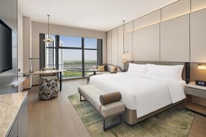 Una habitación de hotel moderna con una cama grande, un escritorio, una silla, un banco, un televisor y vistas a la ciudad.