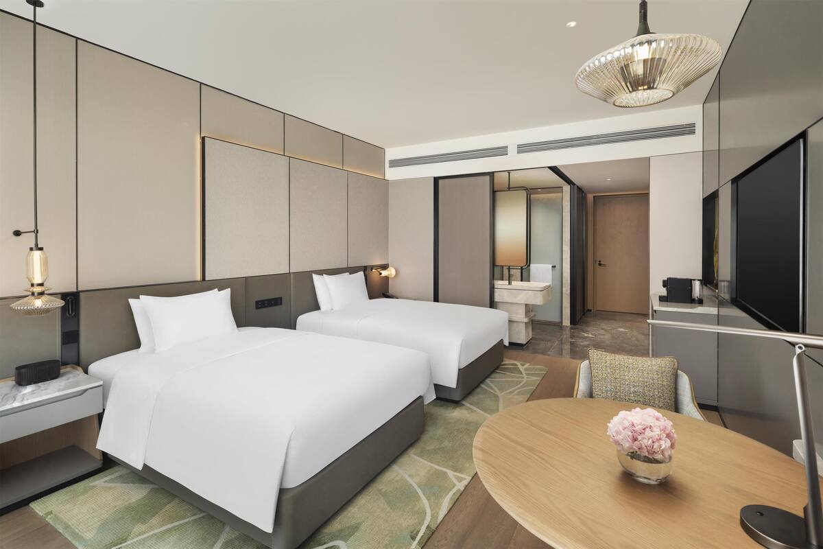 Une chambre d’hôtel moderne avec deux lits, une table à manger, un téléviseur et une salle de bain visible à travers une porte ouverte.