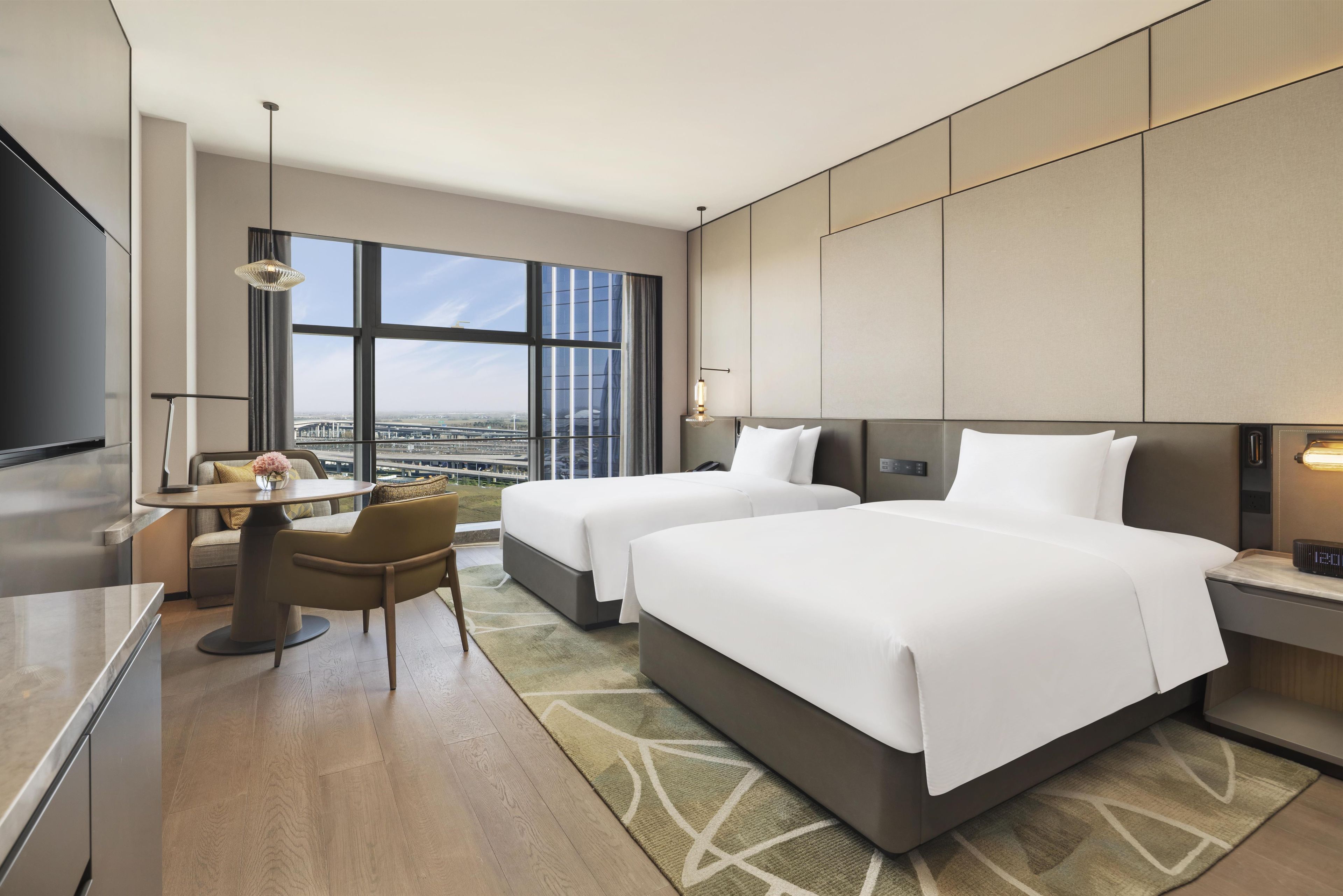 Habitación de hotel con dos camas, zona de comedor, televisión y un amplio ventanal con vistas a la ciudad.