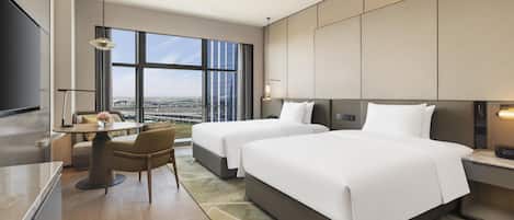A hotel room with two beds, a dining area, a TV, and a large window with a city view.