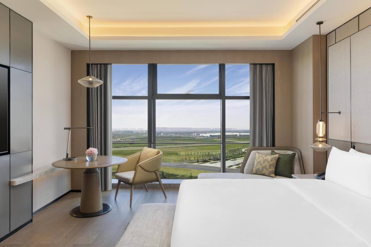Une chambre d’hôtel moderne dotée d’un grand lit, d’une table ronde, de deux chaises, d’une télévision et d’une grande fenêtre donnant sur un paysage verdoyant.