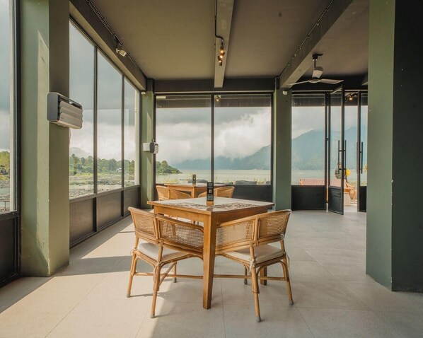 Une pièce spacieuse avec de grandes fenêtres donnant sur un plan d’eau et des montagnes. Au centre, on trouve une table en bois entourée de chaises.