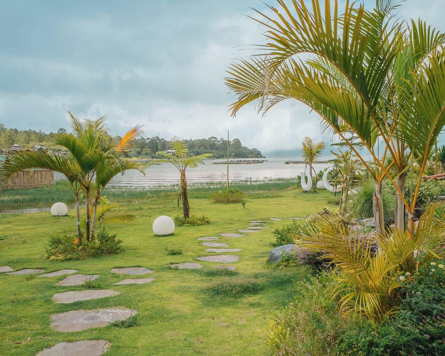 A garden with a stone pathway leading to a body of water, surrounded by greenery and palm trees.