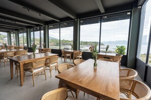 A restaurant with wooden tables and chairs, large windows, and a view of a body of water and mountains.