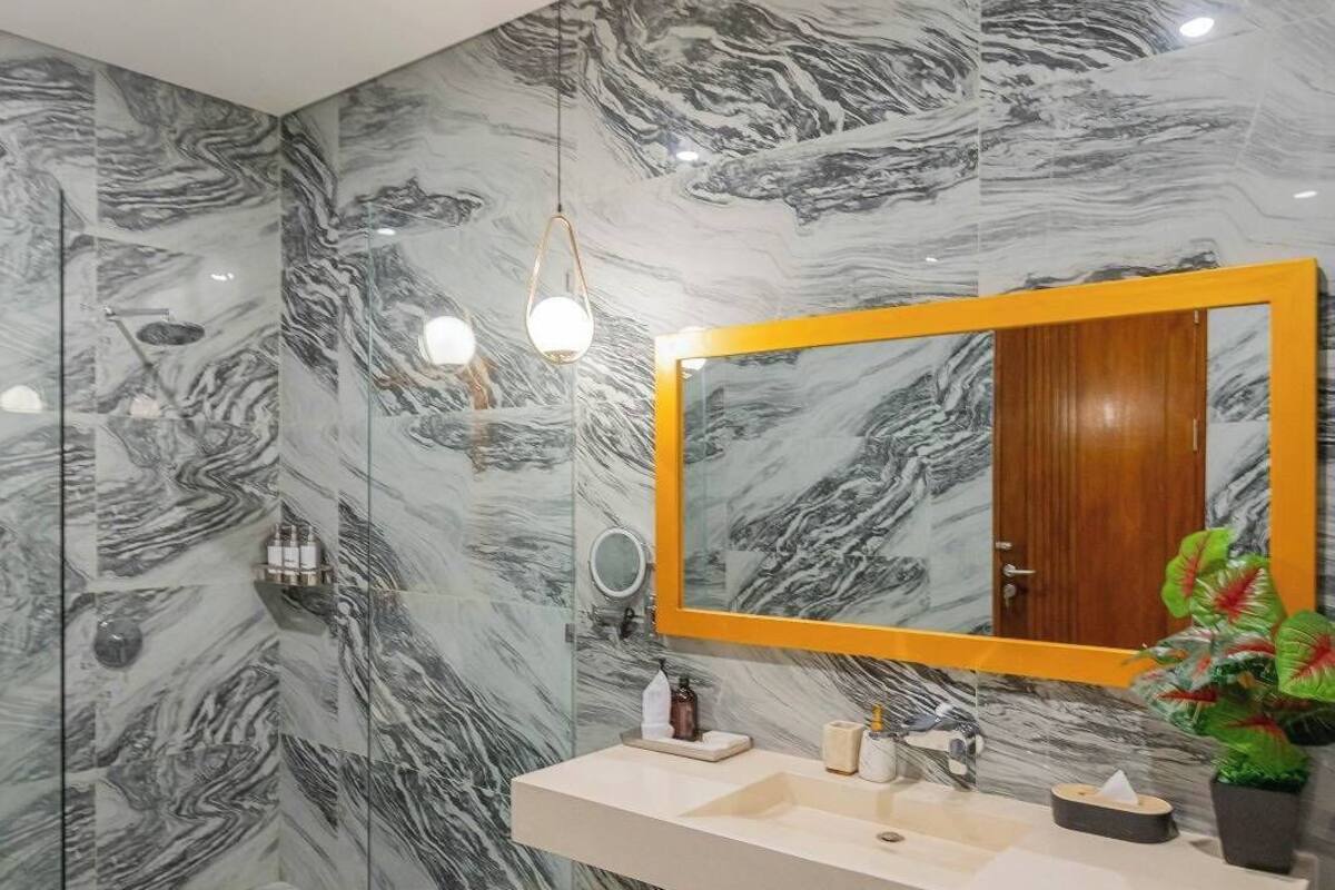 Une salle de bain avec un mur en marbre, un miroir encadré en bois, un lavabo blanc et un luminaire suspendu.