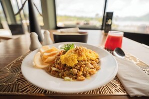 Une assiette de riz frit aux œufs et aux légumes, accompagnée de quelques amuse-gueules frits et d’un verre de boisson rouge, le tout posé sur une table recouverte d’un set de table en fibres tressées.