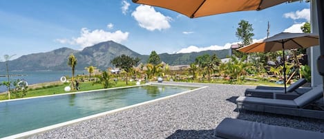 A pool area with lounge chairs and umbrellas, surrounded by a pebble beach and greenery, with a view of a mountain and a body of water.