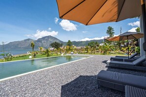 A pool area with lounge chairs and umbrellas, surrounded by a pebble beach and greenery, with a view of a mountain and a body of water.