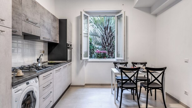A compact kitchen with a built-in washer, a dining area with four chairs, and a window with a view of greenery. - PMT APT (Rome)