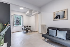 A modern living room with a dark sofa, a dining area with chairs, a large window, and a radiator. - PMT APT (Rome)