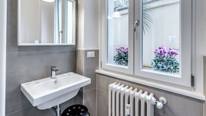 A bathroom with a white sink, a mirror, and a window with a view of flowers. - PMT APT (Rome)