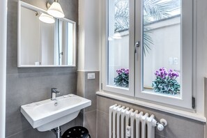 A bathroom with a white sink, a mirror, and a window with a view of purple flowers. - PMT APT (Rome)