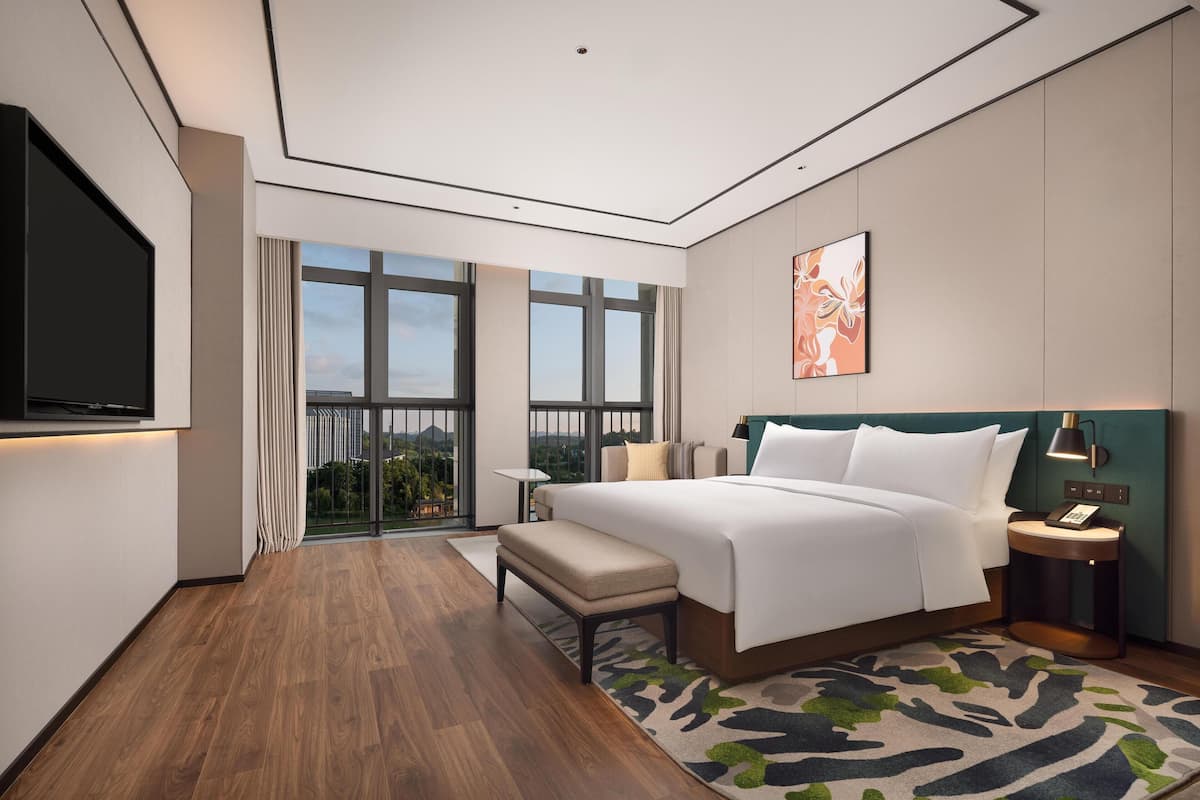 Una moderna camera d'albergo con un ampio letto, una panca, un tavolino, una televisione e vista sulla città attraverso ampie finestre.