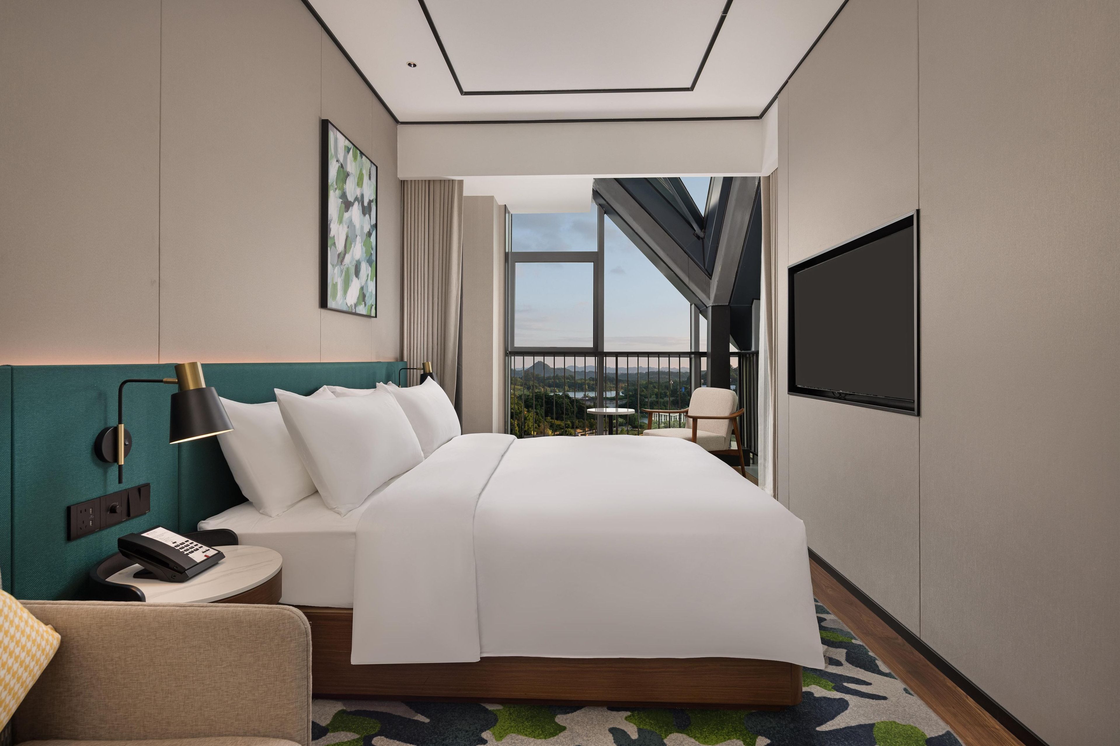 A modern hotel room with a large bed, a desk with a telephone, a chair, a TV, and a balcony with a view.