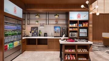 A modern kitchen with a refrigerator, microwave, and coffee machine. There are shelves with snacks and a small table with a fan.