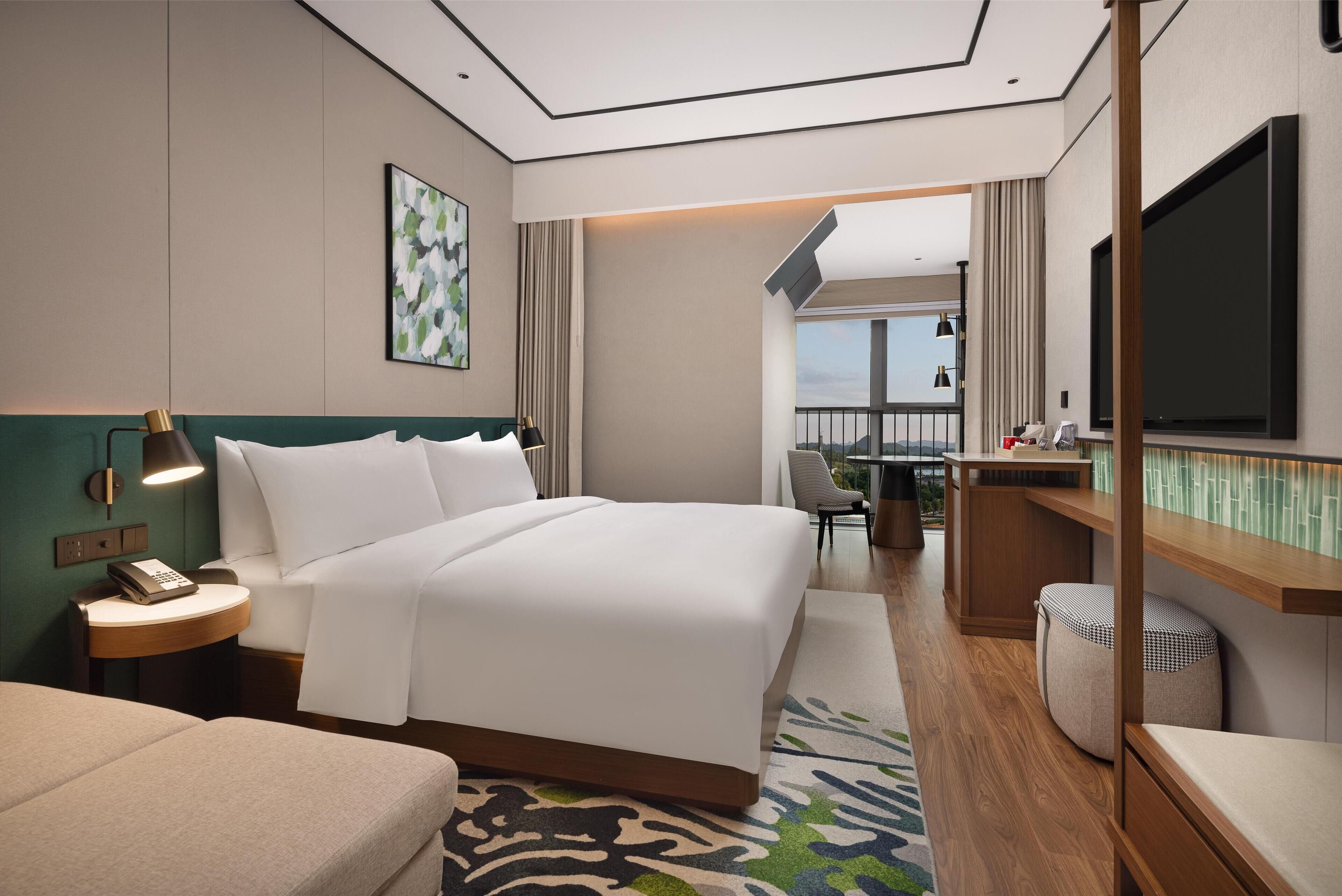 Et moderne hotelværelse med en stor seng, et natbord med lampe, et fjernsyn og udsigt over byen.