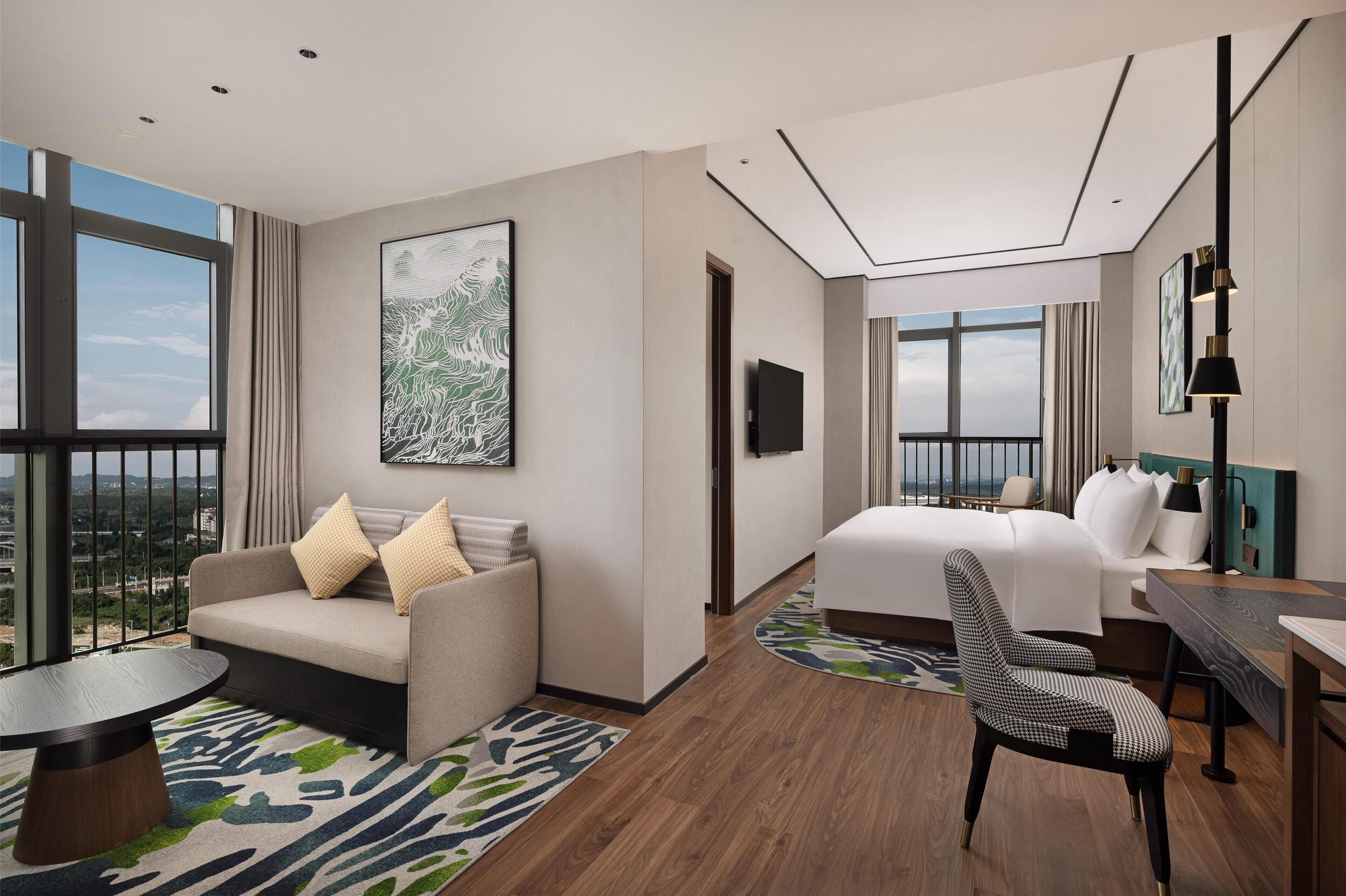 Et moderne hotelværelse med en stor seng, en sofa, et spisebord og en stol. Der er et stort vindue med udsigt over bybilledet.